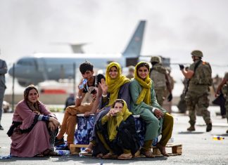 Homem e mulheres afegãs sentados no chão da pista de vôo, avião e exército ao fundo