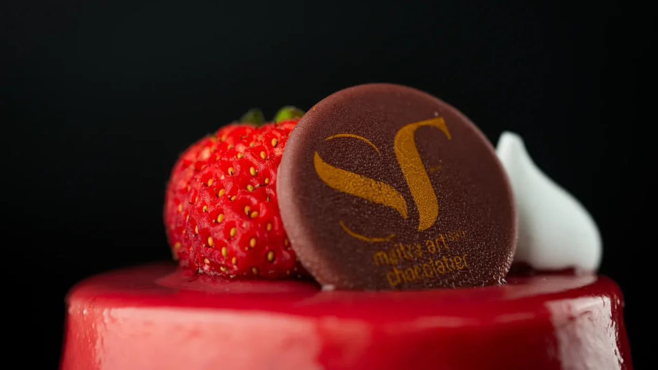 Illustrative photograph. It does not confirm the identity of any person mentioned in the article. Closeup of a red glazed cake topped with fresh strawberry and branded chocolate disc.